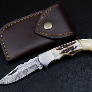 May include: A folding pocket knife with a brown leather sheath. The knife has a stainless steel blade with a damascus pattern and a handle made of white bone with brown accents.