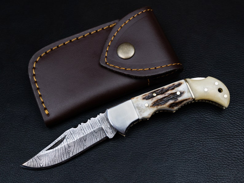 May include: A folding pocket knife with a brown leather sheath. The knife has a stainless steel blade with a damascus pattern and a handle made of white bone with brown accents.