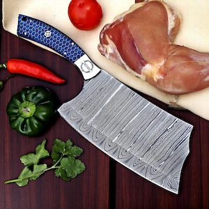 May include: A Damascus steel cleaver with a blue honeycomb patterned handle, alongside fresh ingredients like a tomato, bell pepper, chili pepper, cilantro, and raw chicken. The knife has a silver blade and a decorative handle.