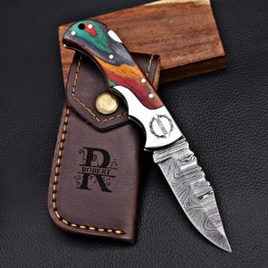 May include: A personalized pocket knife with a colorful wood handle and a Damascus steel blade. The knife is in a brown leather sheath with the name "Robert" engraved on it.