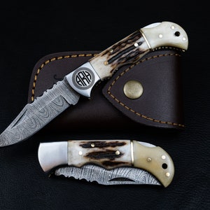 May include: Two pocket knives with a damascus steel blade and a bone handle. The knives are in a brown leather sheath. The top knife has a monogrammed circle on the handle.
