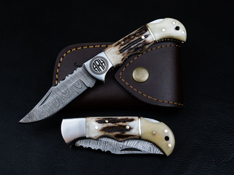 May include: Two pocket knives with a damascus steel blade and a bone handle. The knives are in a brown leather sheath. The top knife has a monogrammed circle on the handle.