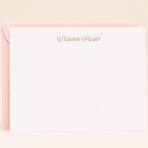 May include: A white stationery card with the name "Elizabeth Torrefiel" printed in gold script, placed on a pink envelope. The stationery set is set against a light beige background.