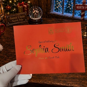May include: A rectangular red envelope featuring gold foil lettering that reads Special delivery for Sophia Smith From the North Pole. The envelope includes a gold stamp graphic in the upper right corner and is held by a white gloved hand.