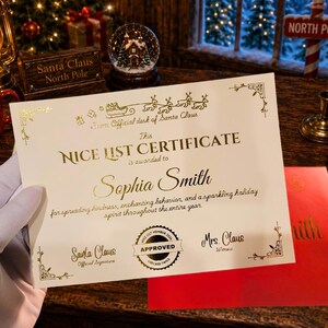 May include: A cream-colored Nice List Certificate with gold foil lettering and decorative accents. The text reads: From Official desk of Santa Claus, This NICE LIST CERTIFICATE is awarded to Sophia Smith, for spreading kindness, enchanting behavior, and a sparkling holiday spirit throughout the entire year. Includes signatures for Santa Claus and Mrs. Claus, plus an Approved seal.
