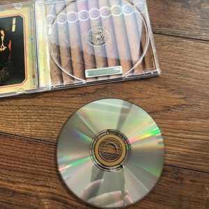 May include: A clear CD case containing cigars, alongside a silver CD. The cigars are brown and neatly arranged. The CD has a reflective surface, showing rainbow hues.