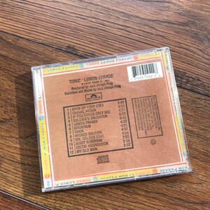 May include: A CD case for the album "Lemon Parade" by Tonic. The case has a brown and tan cardboard backing with the tracklist and album information. The CD case is clear plastic and sits on a wooden surface.