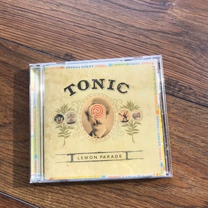 May include: A CD case for Tonic's "Lemon Parade" album. The cover has a vintage aesthetic, with the band's name in bold lettering and a portrait with a swirl over the eyes. The case is transparent, set against a wooden background.