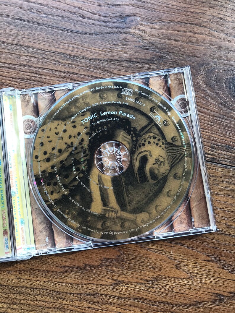 May include: A CD case open on a wooden surface, displaying the disc for Tonic's "Lemon Parade" album. The disc features a sepia-toned design with the band's name and album title. The case is transparent, and the album's tracklist is visible.