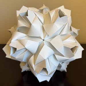 May include: A white origami sphere with a complex, geometric design. The sphere is made of many folded paper triangles and has a textured, three-dimensional appearance.