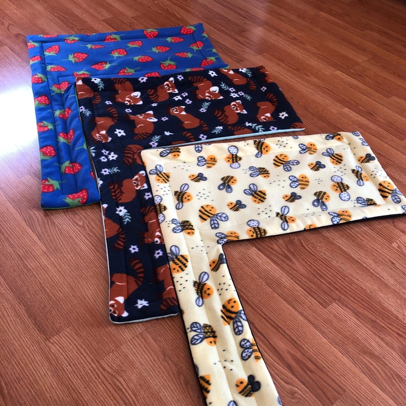 May include: Three rectangular pet beds with different patterns. One is blue with red strawberries, another is navy with red pandas and white flowers, and the third is yellow with bees. The beds are made of soft fabric.
