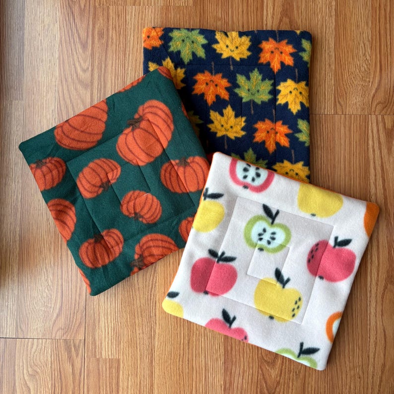 May include: Three square fabric pads featuring seasonal patterns. One pad is dark green with orange pumpkins, one is navy blue with colorful autumn leaves, and one is cream-colored with a repeating pattern of red, yellow, and green apple slices.