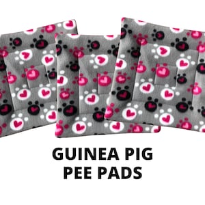 May include: Three gray fleece pee pads for guinea pigs. The pads are patterned with pink hearts and black paw prints on a gray background. The text "GUINEA PIG PEE PADS" is printed below the pads.
