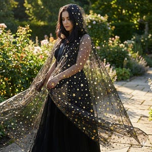 May include: A black hooded cape with a sheer overlay adorned with gold stars. The cape is part of a black dress, creating a celestial and elegant look. The garment is flowing, suggesting movement and a dramatic effect.