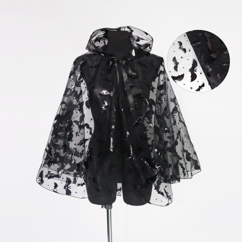May include: Black cape with a hood, adorned with bat and star patterns. The cape features a tie closure at the neck and is displayed on a mannequin. A circular inset shows the bat pattern on white and black backgrounds.