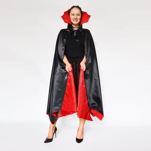 Könnte beinhalten: Ein schwarzer Satinumhang mit rotem Futter und rotem Kragen. Der Umhang ist Teil eines Kostüms. Die Person auf dem Bild trägt ein schwarzes Outfit und schwarze Absätze.