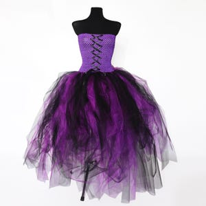 May include: A strapless purple and black tutu dress on a black mannequin. The bodice is solid purple with a criss-cross lace-up design. The skirt has layers of purple and black tulle, creating a full, voluminous effect. A Halloween costume.