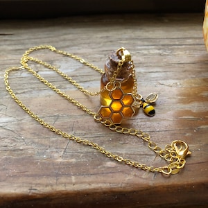 Könnte beinhalten: Goldkette mit einer kleinen Glasflasche gefüllt mit Honig und einem Waben-Anhänger. Die Flasche hat einen Korkverschluss und ist an einer goldenen Kette mit einem Bienen-Anhänger befestigt.