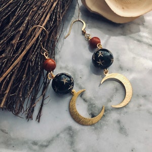 May include: Gold-tone earrings featuring a crescent moon and star design. The earrings have black spherical beads with star cutouts and reddish-brown beads. The earrings are hanging on a gold hook.