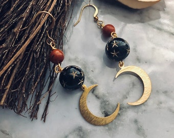 May include: Gold-coloured earrings with a crescent moon and star design. The earrings have black spherical beads with star cutouts and reddish-brown beads. The earrings are suspended from a gold hook.
