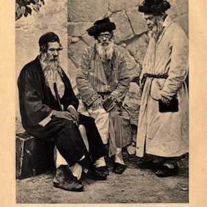 May include: Three men wearing traditional Jewish clothing stand in front of a stone wall. The men are all wearing long beards and hats. The man on the left is wearing a black coat and a white shirt. The man in the middle is wearing a striped robe and a black hat. The man on the right is wearing a white robe and a black hat.