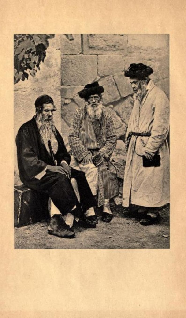 May include: Three men wearing traditional Jewish clothing stand in front of a stone wall. The men are all wearing long beards and hats. The man on the left is wearing a black coat and a white shirt. The man in the middle is wearing a striped robe and a black hat. The man on the right is wearing a white robe and a black hat.