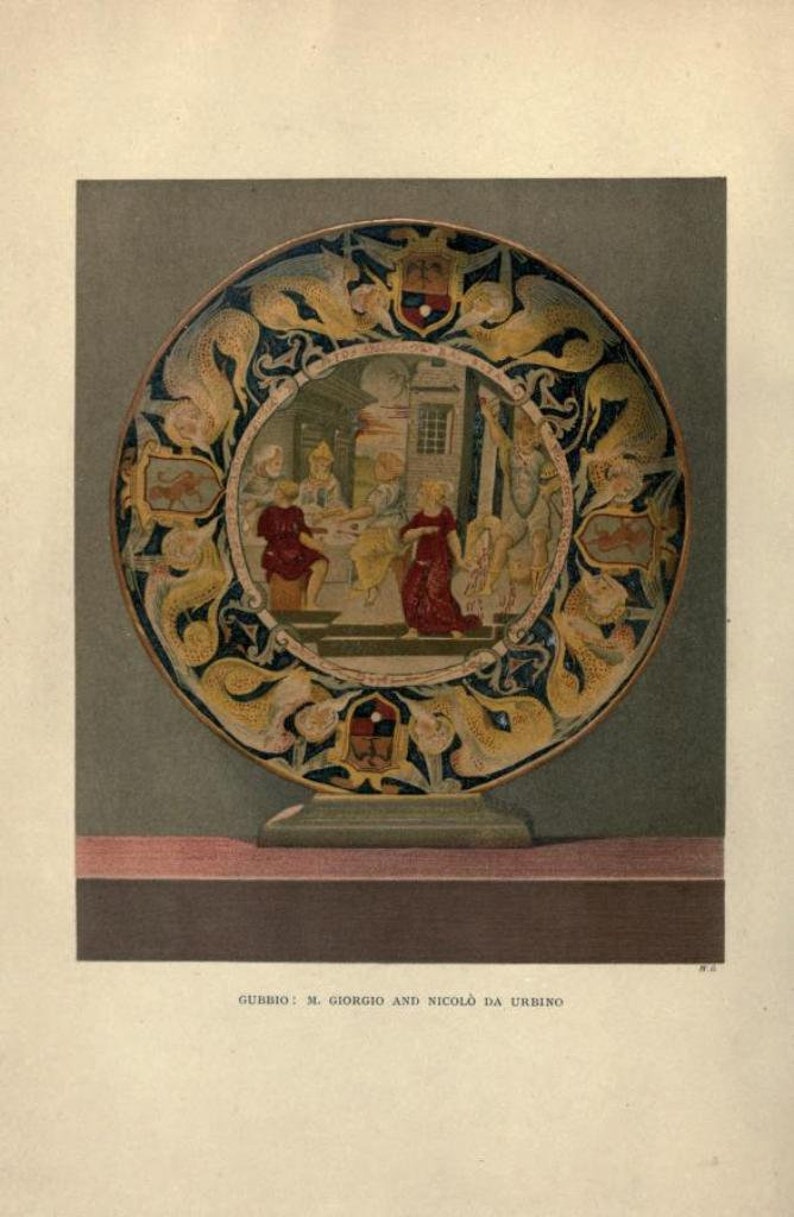 Op de afbeelding: Een rond keramiek bord met een kleurrijk ontwerp. Het bord heeft een centraal tafereel van mensen die rond een tafel zijn verzameld, omringd door een rand van gouden en blauwe draken. De tekst "GUBBIO: M. GIORGIO AND NICOL&Ograve; DA URBINO" is onder het bord gedrukt.
