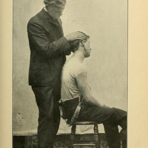 May include: A black and white photo of a person demonstrating a technique for locating the transverse process of the atlas bone. The person is wearing a suit and is touching the head of the other person, who is sitting on a chair.