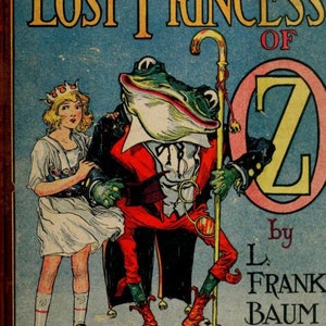以下が含まれることがあります： L. フランク・バウムによる「オズの迷子の王女」の表紙イラスト。表紙には、赤いスーツと白いシャツを着て杖を持った緑色のカエルが描かれている。白いドレスと王冠をつけた若い女の子がカエルの隣に立っている。背景は青で、ピンクの円の中に大きな金の文字「Z」がある。