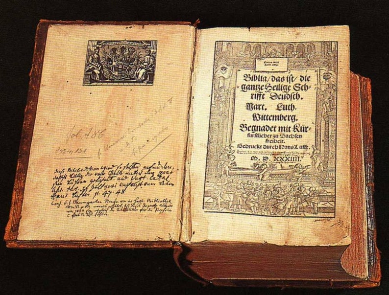 May include: An antique, leather-bound book with a worn, aged appearance. The cover is brown with a faded, gold-colored design. The book is open to a page with a black and white illustration and text in German. The text reads "Biblia/das ist die ganze Heilige Schriffe Seudsch. Warr. Luth. Wittemberg. Begnadet mit K&uuml;r fur lieber zu Bacbfen freibeir. Bedruckt durch Pano uffy. .іїааа.."