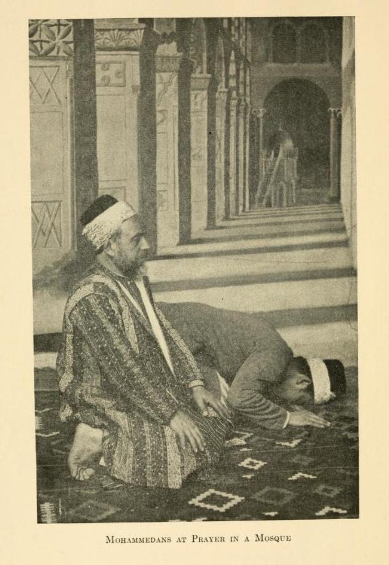 May include: Two men are kneeling in prayer in a mosque. The man in the foreground is wearing a white turban and a long robe. The man in the background is wearing a white turban and a long robe. The mosque is decorated with intricate tile work.