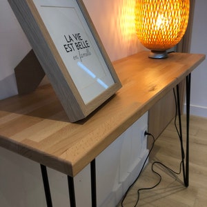 Peut inclure: Une console en bois avec des pieds en épingle à cheveux noirs. Un cadre avec l'inscription "La vie est belle en famille" est posé sur la table. Une lampe en osier tressé avec une lumière chaude est sur la table.