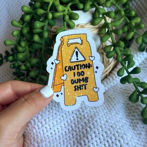 May include: A yellow sticker in the shape of a caution sign with the text "CAUTION: I DO DUMB SH!T". The sticker has a white border, hearts, and sparkles. The sticker is held by a hand with white nail polish.