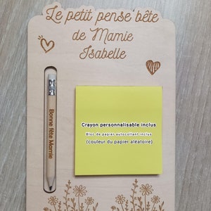 Puede incluir: Un tablero de notas de madera con el texto "Le petit pense bete de Mamie Isabelle" y un pequeño corazón. El tablero tiene una ranura para un lápiz e incluye un bloc de notas adhesivas amarillo y un lápiz. El tablero está decorado con un diseño floral.
