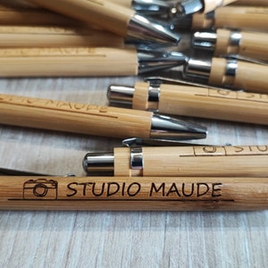 Puede incluir: Primer plano de un montón de bolígrafos de bambú con detalles plateados. Los bolígrafos están grabados con el texto "Studio Maude" y un icono de cámara.