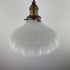 May include: A pendant light fixture featuring a translucent, fluted white glass shade with a scalloped edge. The shade is attached to a metallic, antique-brass finish socket and hardware, suspended by a brown fabric-covered electrical cord.
