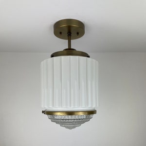 May include: A white and brass ceiling light fixture. The fixture has a cylindrical white glass shade with vertical ridges, a brass-colored metal base, and a clear, textured glass bottom section. The light is suspended from a brass-colored ceiling mount.