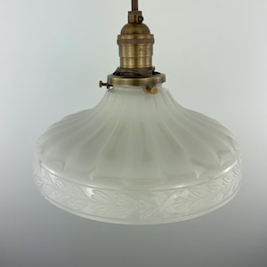 May include: A vintage-style pendant light fixture with a frosted glass shade. The shade has a fluted design and a decorative leaf pattern around the bottom. The fixture includes a brass-colored socket and a brown cord.