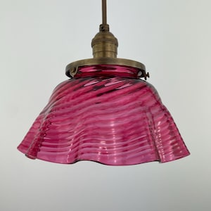 May include: A vintage pendant light with a pink glass shade. The shade has a wavy design and is suspended from a brass-toned fixture and a brown cord. The light fixture is designed for overhead illumination. The shade's diameter is approximately 20 cm.