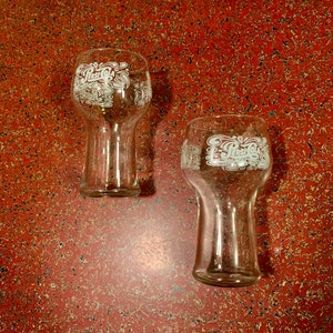May include: Two clear glass Pepsi glasses with the Pepsi logo printed in white on the front. The glasses are sitting on a red speckled surface.