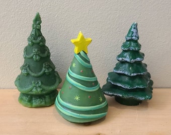 Puede incluir: Tres figuras de árboles de Navidad verdes. Uno tiene una estrella amarilla y rayas verdes y blancas. Los otros dos son de color verde liso con diseños detallados. Las figuras están sobre una superficie de madera.