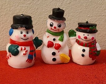 May include: Three small snowman figurines with black top hats, white bodies, and colorful scarves. The snowman on the left has a red and white striped scarf, the middle snowman has a green scarf, and the snowman on the right has a green and red striped scarf.