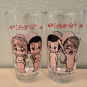 May include: Two clear glass tumblers with a cartoon illustration of two figures holding flowers. The text "Love is..." is above the figures in pink, with additional text at the base of each glass.