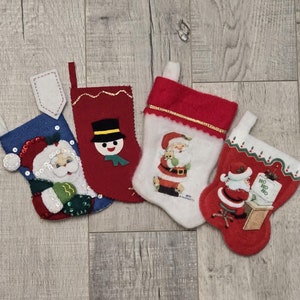 May include: Four miniature Christmas stockings in red, white, and blue. Each stocking features a different festive design, including Santa Claus, a snowman, and the words "HO HO HO". These decorative stockings are perfect for holiday decor.