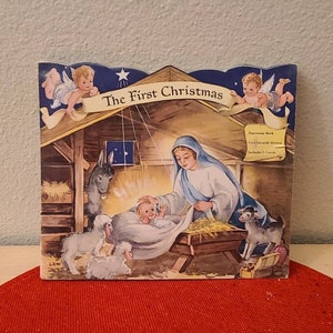 May include: A vintage children's book titled "The First Christmas" with a colorful illustration of the nativity scene. The cover features Mary holding baby Jesus in a manger, with a donkey, sheep, and angels.