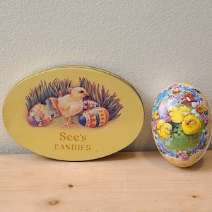 May include: Two oval-shaped tins with Easter-themed designs. The larger tin is yellow with a chick and eggs, and the text "See's CANDIES". The smaller tin is white with a blue border and a cluster of yellow chicks in a nest.