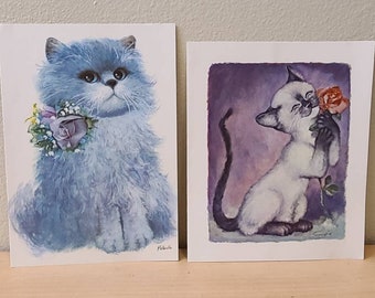 May include: Two cat art prints. One features a blue Persian cat with a floral collar. The other depicts a Siamese cat holding a rose. Both prints have a vintage, illustrative style and are displayed on a wooden surface.