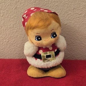 May include: A plush toy elf with a red and white polka dot hat, a white fur trim coat, and a black belt with a gold buckle. The elf has a smiling face with blue eyes and rosy cheeks.