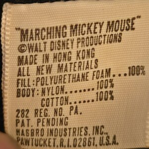 Peut inclure: Gros plan sur une &eacute;tiquette rectangulaire blanc cass&eacute; avec du texte noir. L'&eacute;tiquette indique "MARCHING MICKEY MOUSE" et comprend des d&eacute;tails sur Walt Disney Productions, le lieu de fabrication, les mat&eacute;riaux et une adresse de Hasbro Industries.