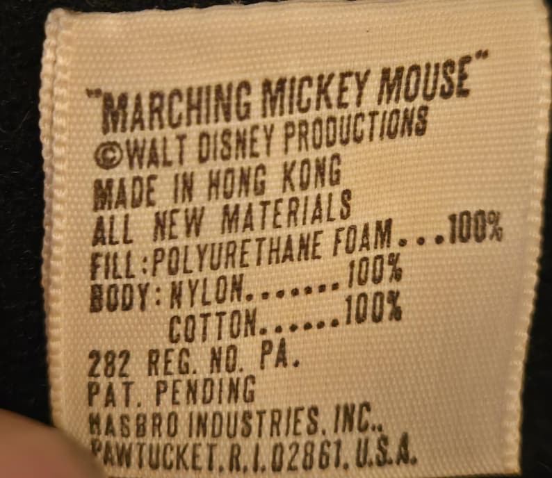 Peut inclure: Gros plan sur une &eacute;tiquette rectangulaire blanc cass&eacute; avec du texte noir. L'&eacute;tiquette indique "MARCHING MICKEY MOUSE" et comprend des d&eacute;tails sur Walt Disney Productions, le lieu de fabrication, les mat&eacute;riaux et une adresse de Hasbro Industries.